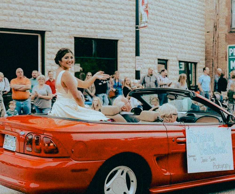 Jordan Mn Heimetfest celebration shows a scene of many people standing in the foreground as a parade of cars flow by. The main car shows a girl sitting in a dress waving as two people sit up front.