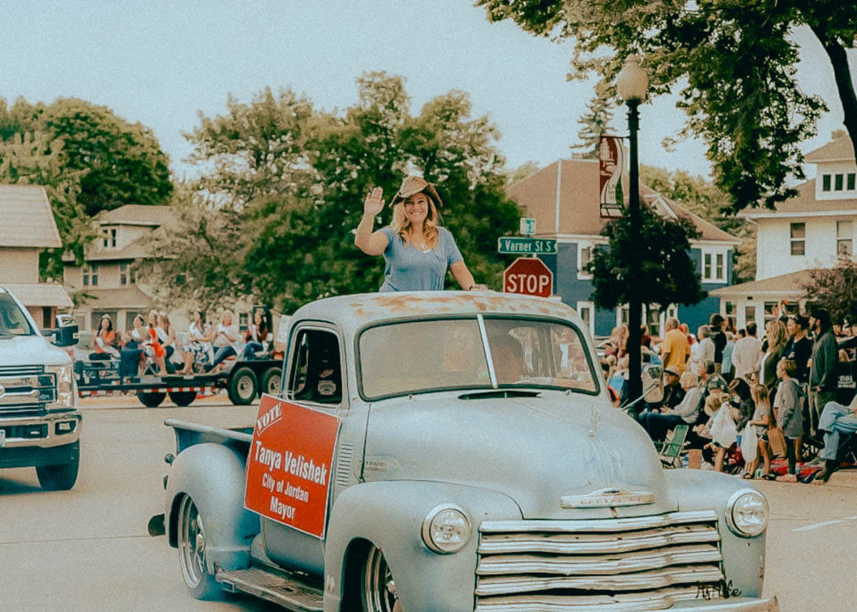 Tanya Velishek rides on the back of a old vintage truck with a mayor sign on the side during the Heimefest parade. She is wearing a cowboy hat and waving as she rides past.